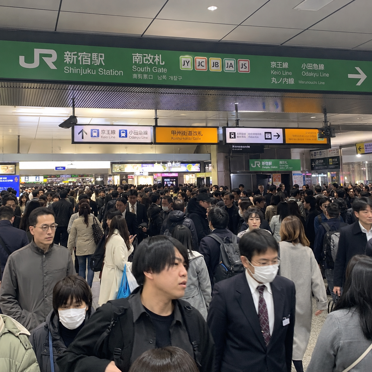 A crowded Shinjuku Station platform in Japan, with many passengers walking under green JR station signage and overhead platform displays.
